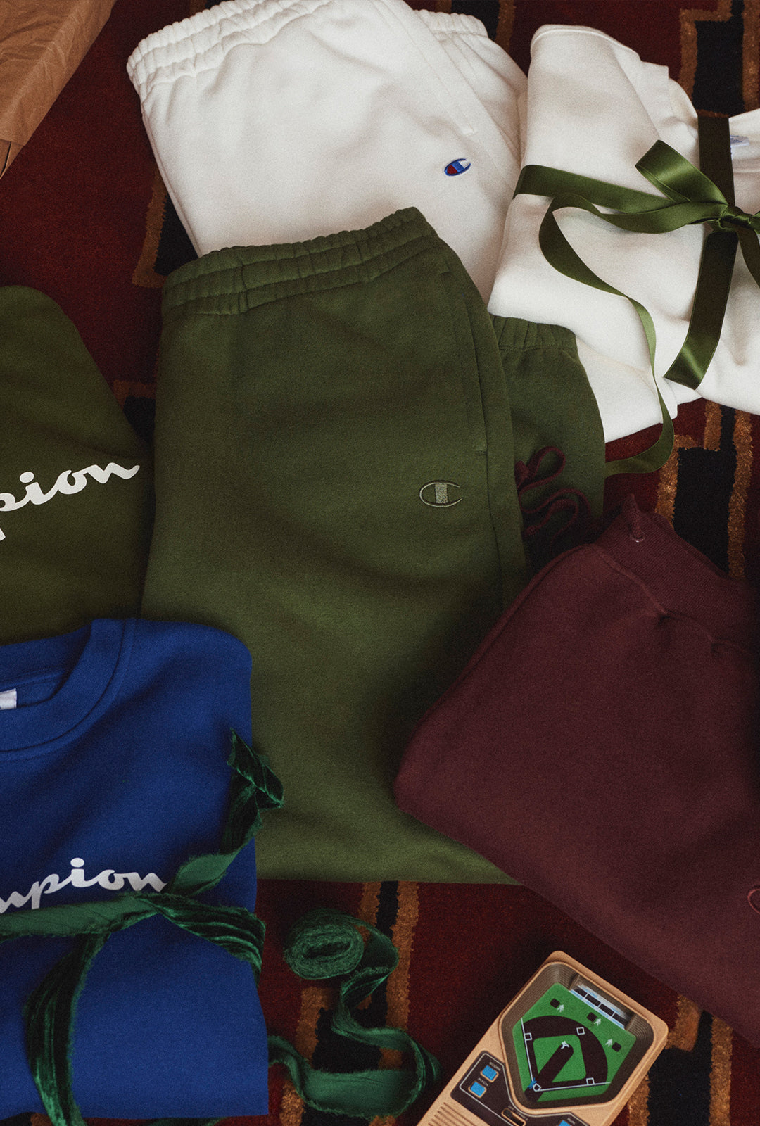 A styled flat-lay of multiple Champion sweatpants and sweatshirts in different colors, including white, olive green, blue, and burgundy, arranged on a patterned red rug with a small baseball card.