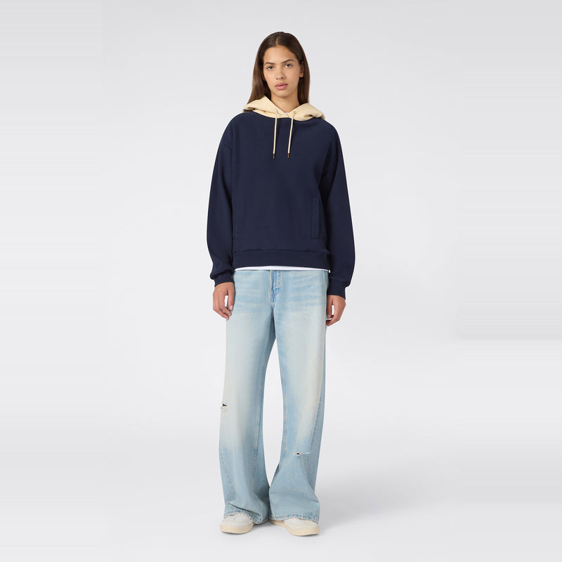 A person stands facing forward, wearing a navy Champion Women’s Knitted Hooded Sweatshirt layered over a beige hood, paired with light blue wide-leg jeans and white sneakers, against a plain light gray background.
