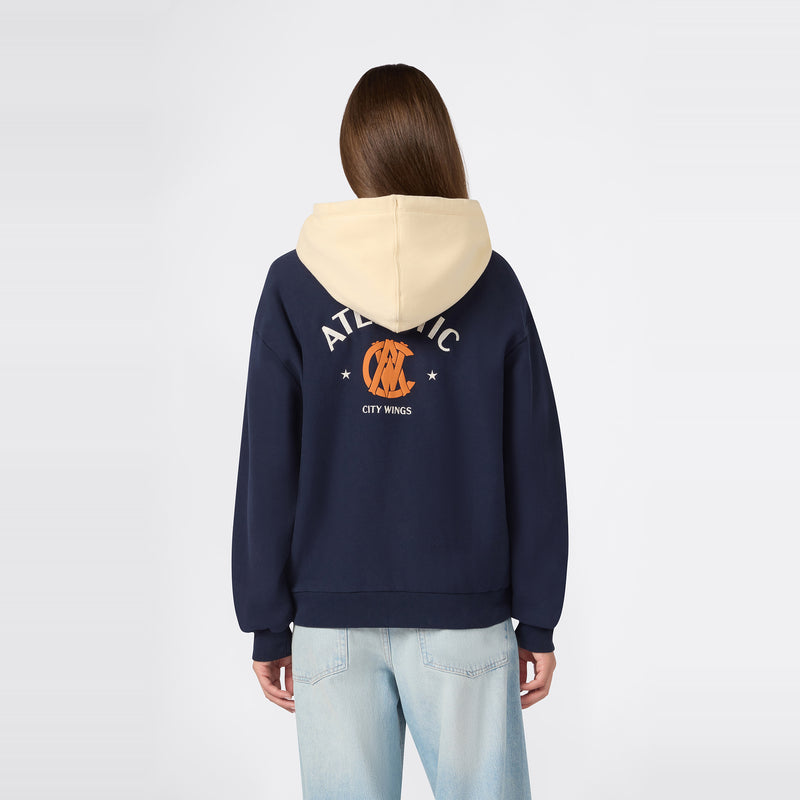 A person wearing the Champion Women’s Knitted Hooded Sweatshirt in navy, featuring a beige hood, “ATLANTIC” text, an orange graphic on the front, and “CITY WINGS” printed on the back, paired with light blue jeans, stands facing away from the camera against a plain background.