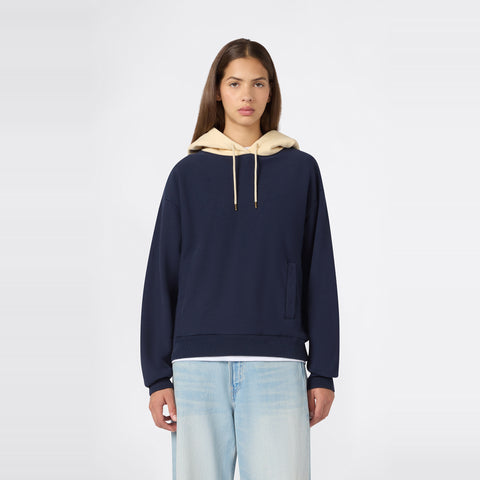 A person wearing the Champion Women’s Knitted Hooded Sweatshirt in navy blue, featuring a beige hood and heavyweight fleece construction, paired with light blue jeans, stands facing forward against a plain white background.