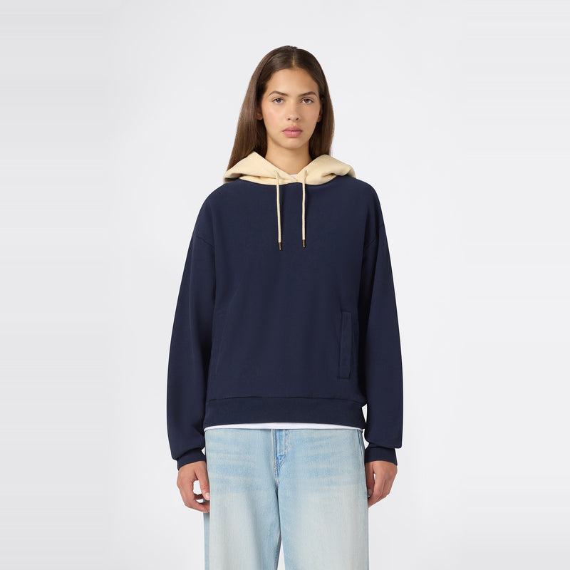 A person wearing the Champion Women’s Knitted Hooded Sweatshirt in navy blue, featuring a beige hood and heavyweight fleece construction, paired with light blue jeans, stands facing forward against a plain white background.