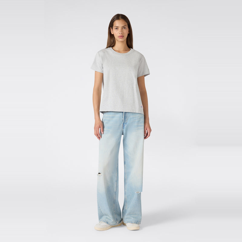 A woman stands facing forward against a plain white background, wearing the Champion Women’s Knitted Crewneck T-Shirt in grey with a relaxed fit, paired with light blue wide-leg jeans featuring subtle distressing and white sneakers.