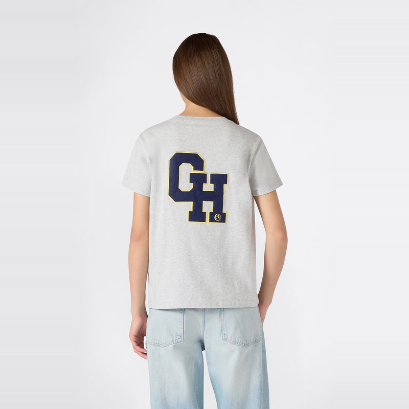 A woman with long brown hair is wearing a relaxed fit light gray Champion Women’s Knitted Crewneck T-Shirt, featuring large blue and yellow “CH” letters on the back, paired with light blue jeans, while standing against a plain white background.