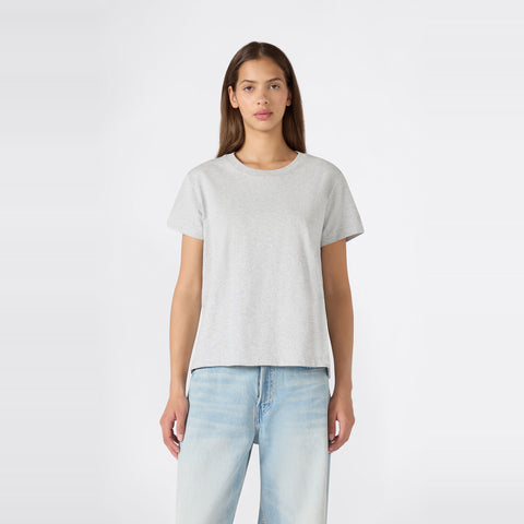 A person with long brown hair stands facing forward against a plain white background, wearing a relaxed fit Champion Women’s Knitted Crewneck T-Shirt in plain light gray paired with light blue jeans.