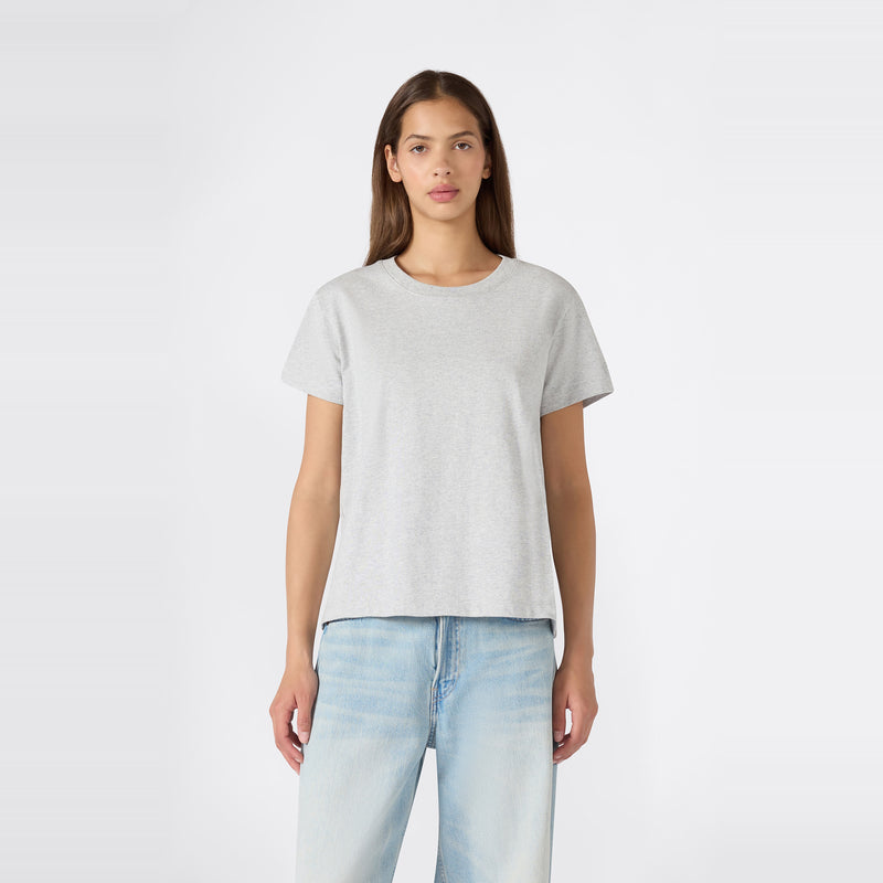 A person with long brown hair stands facing forward against a plain white background, wearing a relaxed fit Champion Women’s Knitted Crewneck T-Shirt in plain light gray paired with light blue jeans.