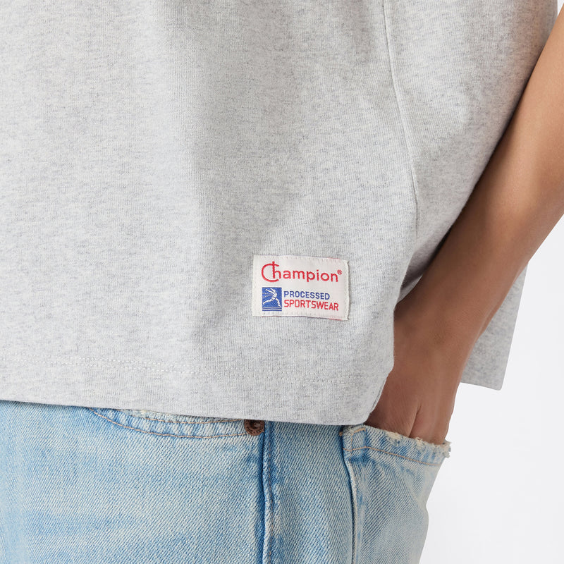 A person wearing a light gray Champion Women’s Knitted Crewneck T-Shirt and light blue jeans, with one hand in their pocket. The relaxed fit of the t-shirt enhances the casual look, and the Champion logo patch is visible near the hem.