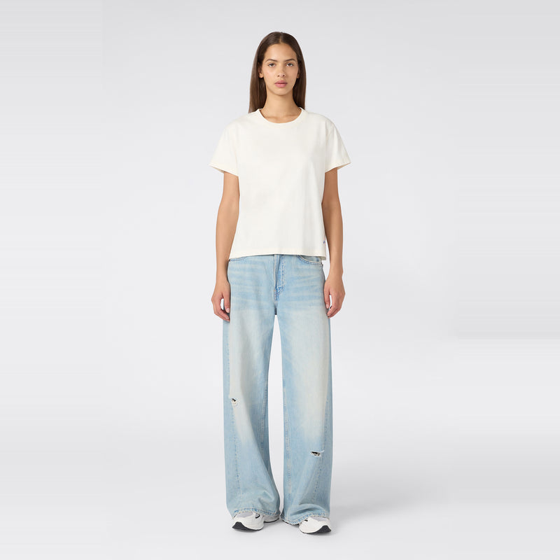 A woman faces forward wearing a Champion Women’s Knitted Crewneck T-Shirt in plain white, paired with light blue wide-leg ripped jeans and white sneakers, set against a simple light background.