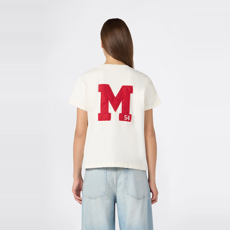A woman with long brown hair is wearing the Champion Women’s Knitted Crewneck T-Shirt in white, featuring a bold red “M” and a small “54” on the back, paired with relaxed fit light blue jeans, standing against a plain white background.