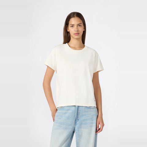 A woman with straight brown hair is wearing a relaxed fit Champion Women’s Knitted Crewneck T-Shirt in plain white paired with light blue jeans, standing against a light gray background with one hand in her pocket.