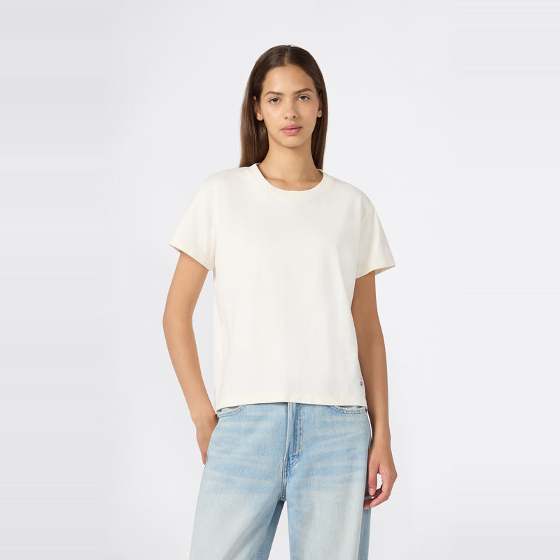 A woman with straight brown hair is wearing a relaxed fit Champion Women’s Knitted Crewneck T-Shirt in plain white paired with light blue jeans, standing against a light gray background with one hand in her pocket.
