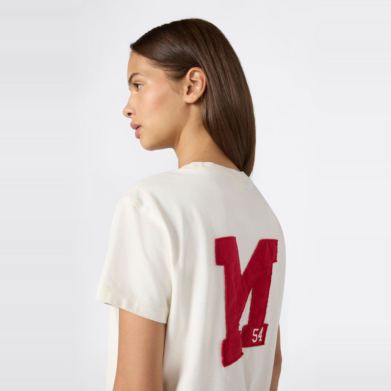A woman with straight brown hair models the Champion Women’s Knitted Crewneck T-Shirt, featuring a relaxed fit. The shirt is styled with a bold red letter M and the number 54 on the back. She stands against a plain, light background, highlighting this Champion look inspired by their iconic Reverse Weave collection.