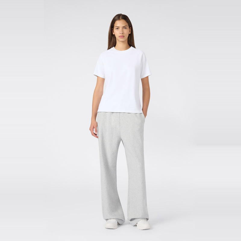 A person stands against a plain light background, wearing a white short-sleeve t-shirt, Champion Women’s Knitted Flare Pants in light gray, and white shoes. Their hands are in their pockets, and they have straight brown hair.