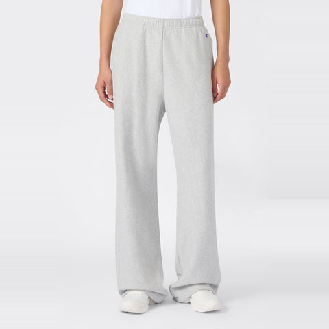 A person wearing Champion Women’s Knitted Flare Pants in a loose-fit, light gray style, paired with a white t-shirt and white sneakers, stands against a plain white background. The heavyweight fleece fabric of these flared pants adds an elevated touch to this classic women’s sportswear look.