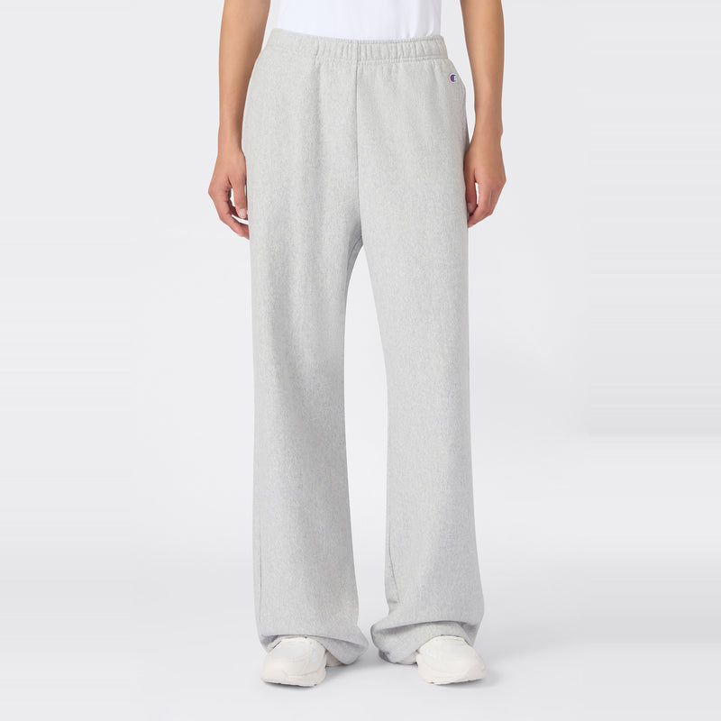 A person wearing Champion Women’s Knitted Flare Pants in a loose-fit, light gray style, paired with a white t-shirt and white sneakers, stands against a plain white background. The heavyweight fleece fabric of these flared pants adds an elevated touch to this classic women’s sportswear look.