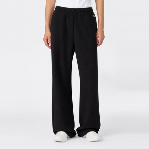 A person wearing loose black Champion Women’s Knitted Flare Pants and white sneakers stands against a plain white background. Only the lower half of the body is visible, with hands relaxed at the sides, highlighting a laid-back approach to women’s sportswear.