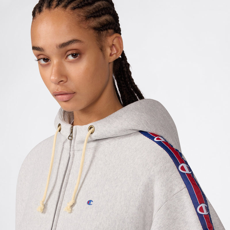 A person with braided hair wears the Women’s Icon Hooded Full Zip Sweatshirt by Champion in light gray, featuring blue and red logo stripes on the sleeves, looking slightly to the side against a plain background. The heavyweight fleece construction of this hoodie provides both comfort and durability, making it a stylish choice.