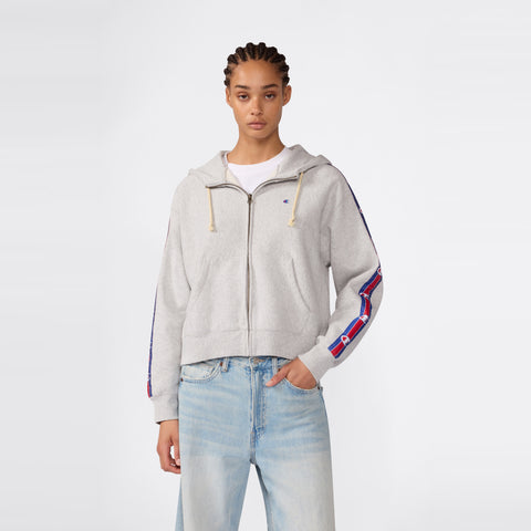 A woman wearing the Champion Women’s Icon Hooded Full Zip Sweatshirt in light gray, paired with blue jeans, stands facing forward against a plain white background.