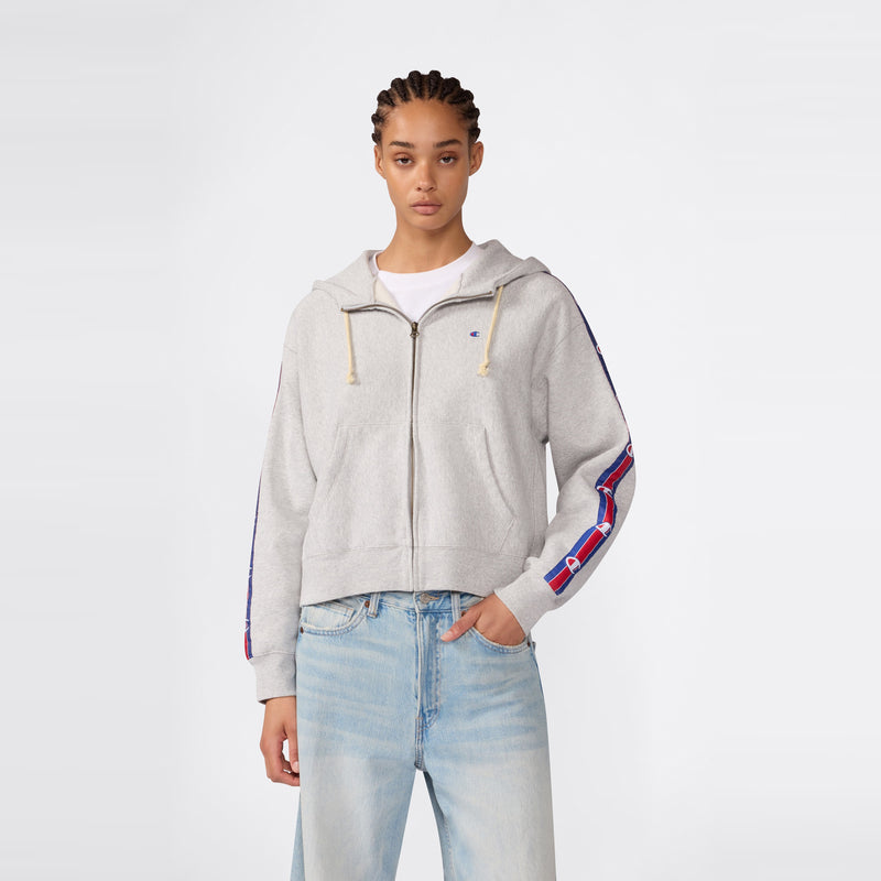 A woman wearing the Champion Women’s Icon Hooded Full Zip Sweatshirt in light gray, paired with blue jeans, stands facing forward against a plain white background.