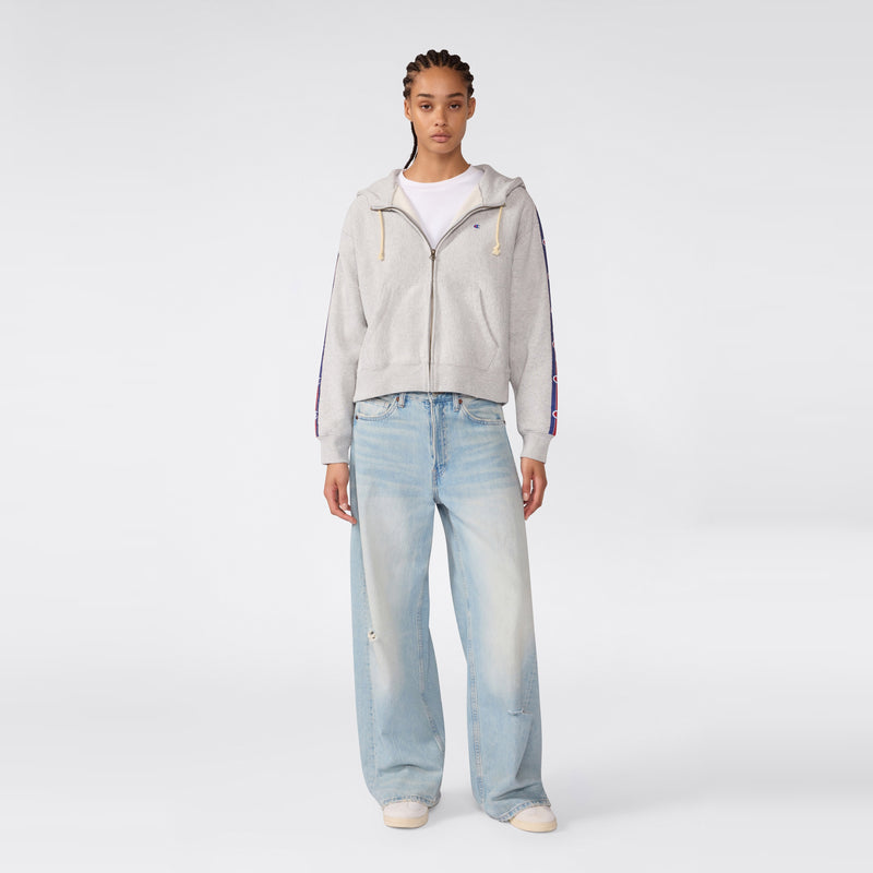 A person stands facing forward, wearing a Champion Women’s Icon Hooded Full Zip Sweatshirt in gray over a white shirt and loose-fitting light blue jeans, paired with white sneakers, against a plain light gray background.
