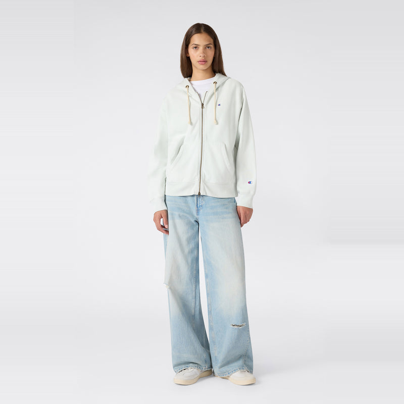 A person stands facing forward against a plain light background, wearing a white Champion Women’s Knitted Hooded Full Zip Sweatshirt zipped up over a white t-shirt, paired with light blue wide-leg jeans and white sneakers.