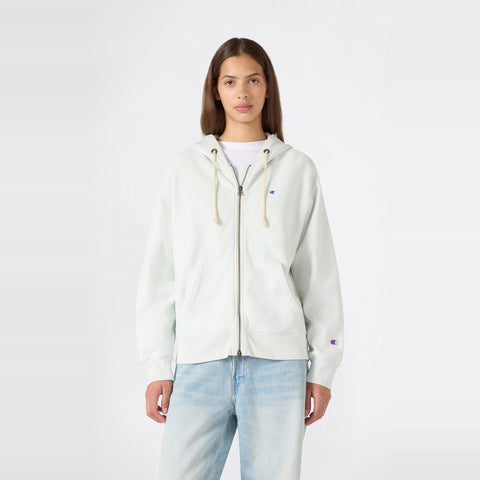 A person stands facing forward, wearing a Champion Women’s Knitted Hooded Full Zip Sweatshirt in a light color over a white shirt and paired with light blue jeans, against a plain white background.