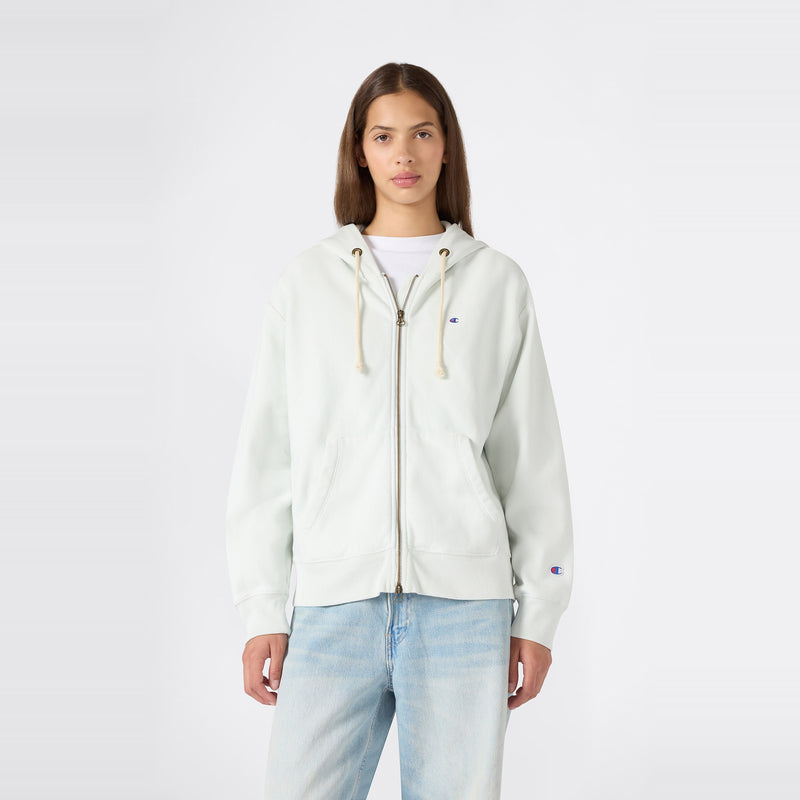 A person stands facing forward, wearing a Champion Women’s Knitted Hooded Full Zip Sweatshirt in a light color over a white shirt and paired with light blue jeans, against a plain white background.
