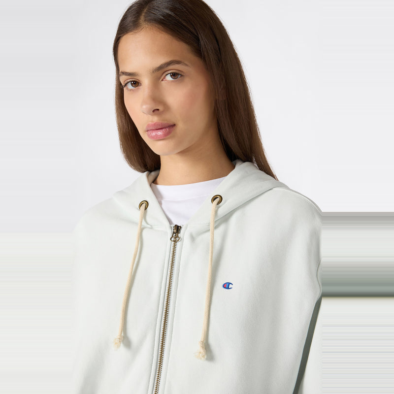 A woman with long brown hair wears the Champion Women’s Knitted Hooded Full Zip Sweatshirt, which features tan drawstrings and a small blue Champion logo, layered over a white shirt against a plain background.
