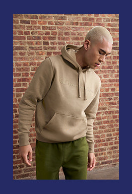 Person wearing a beige hoodie with a brand logo against a brick wall