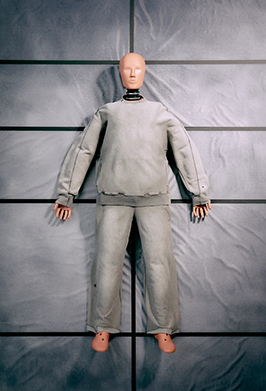 A life-sized mannequin or doll wearing a loose gray long-sleeve top and matching pants, standing against a gray paneled backdrop.