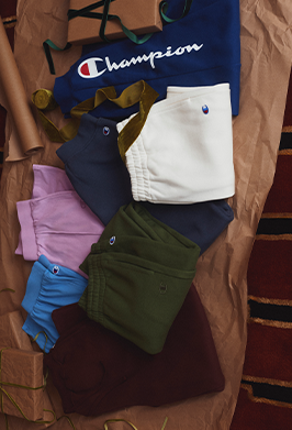 A flat-lay arrangement of multiple folded Champion garments on brown paper, including a navy sweatshirt with the Champion logo, white shorts, navy pants, green pants, a light purple top, blue shorts, and a dark burgundy top.