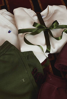 A folded white sweatshirt tied with a dark green ribbon, layered over matching green sweatpants and burgundy sweatpants, with Champion logos visible.