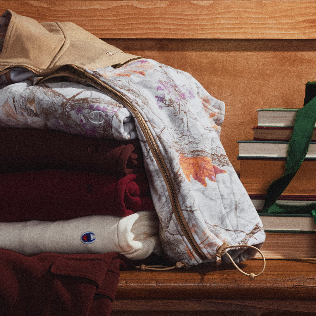 A close-up of a zippered bag with a woodland camouflage print, resting on top of folded garments on a wooden surface.