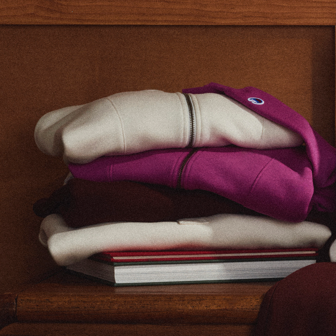 A stack of folded sweatshirts in cream, magenta, and dark brown, resting on books against a wooden background. The top sweatshirt features a small embroidered Champion logo.