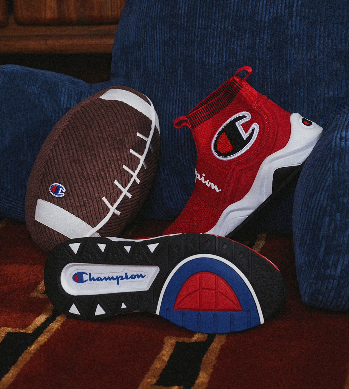 A pair of red, white, and black Champion high-top sneakers arranged on a patterned rug in front of a blue textured armchair, with a brown football prop behind them.
