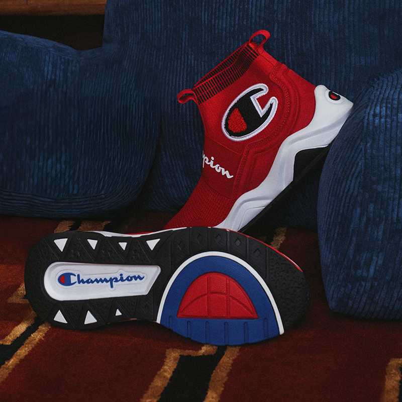 A pair of Champion high-top sneakers in red, white, and blue, with one shoe showing the red knit upper and the other showing the black, white, and blue outsole, set against a dark blue textured couch.