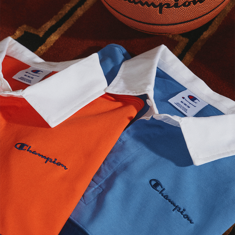 Two Champion short-sleeve collared shirts, one orange and one light blue, arranged on a patterned rug with a basketball partially visible at the top.
