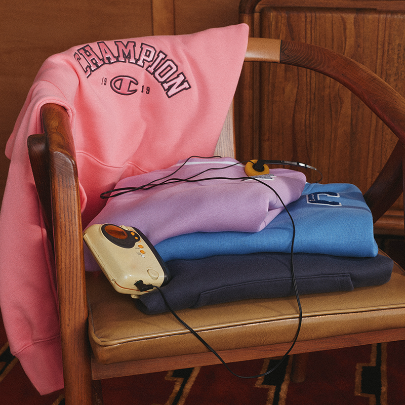 A pink Champion sweatshirt draped over a wooden chair, with folded sweatshirts in lavender, blue, and navy stacked on the seat. A cream handheld device with black cords rests on top.