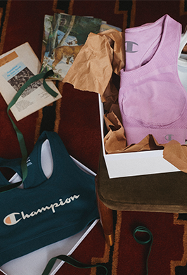 A styled flat-lay of Champion athletic apparel and accessories on a patterned rug, including a dark teal cropped tank top with the Champion logo, a light purple sleeveless top, brown tissue paper, and a white shoebox.