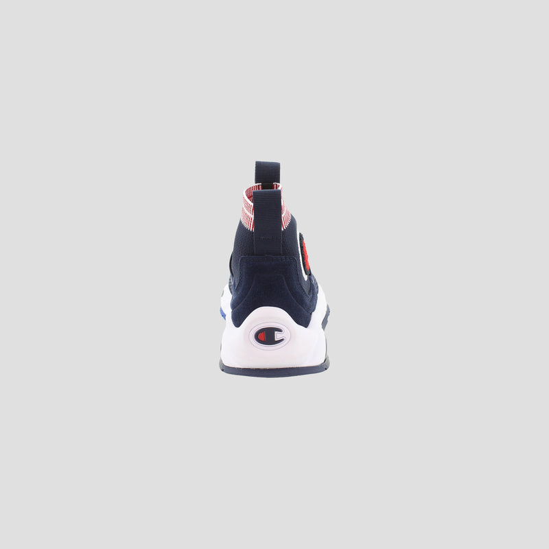 The Champion Kids' Rally Pro slip-on shoe is shown from the back, highlighting its navy upper, white sole, Champion logo on the heel, and a red and white striped pull tab. The background is plain light gray.