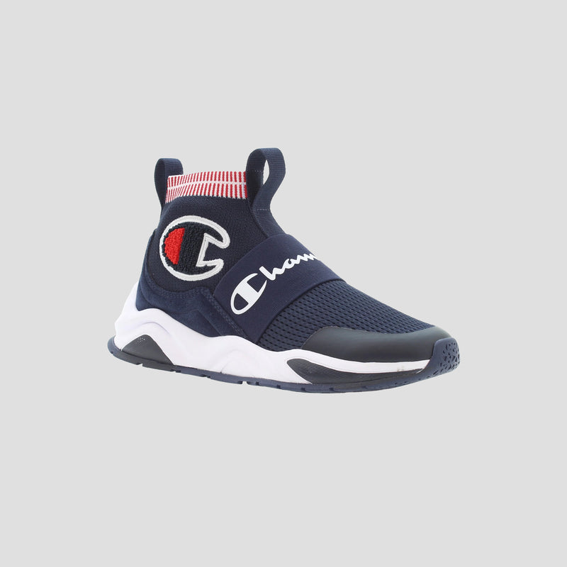 The Kids' Rally Pro by Champion is a navy slip-on shoe with a white sole, red and white striped ankle, and prominent Champion logo on the side and strap, shown against a light gray background.