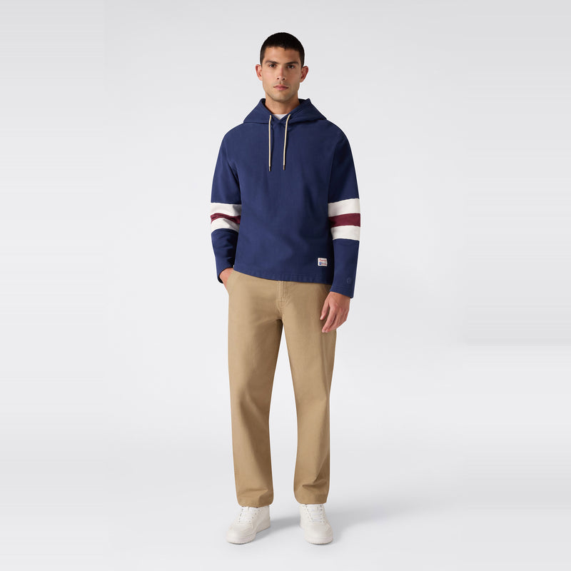 A man stands facing forward, wearing a Champion Men’s Knitted Hooded Sweatshirt in navy with white and maroon stripes on the sleeves, khaki pants, and white sneakers against a plain light background.