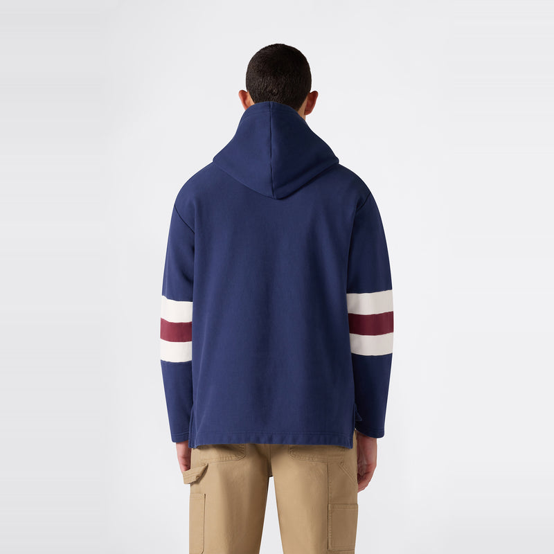A person stands facing away, wearing the Champion Men’s Knitted Hooded Sweatshirt in navy blue with white and red stripes on the sleeves, paired with beige cargo pants, against a plain white background. This hoodie features heavyweight fleece for added warmth and comfort.