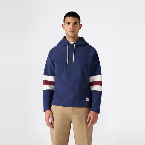 A man stands facing forward against a plain light background, wearing a navy Champion Men’s Knitted Hooded Sweatshirt with white and maroon stripes on the sleeves, paired with tan pants.