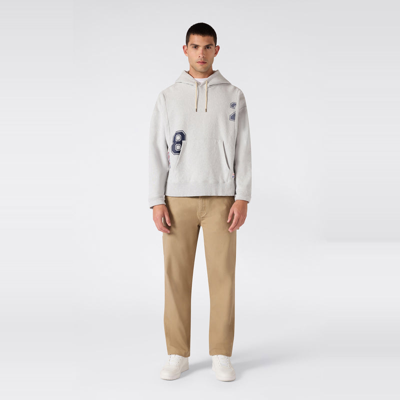 A person stands facing forward, wearing a light gray Champion Men’s Knitted Hooded Sweatshirt (College Graphic), crafted from soft fleece with patches, paired with tan pants and white sneakers, on a plain white background.