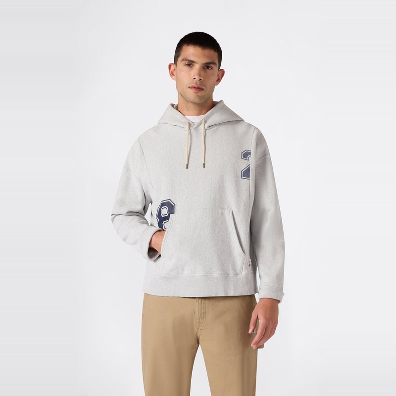 A man wearing a light gray Champion Men’s Knitted Hooded Sweatshirt (College Graphic) and tan pants stands against a plain white background, facing forward with one hand in his pocket, exemplifying classic college-inspired style.