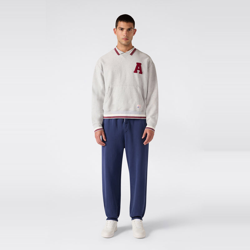 A person stands facing forward, wearing the Champion Men’s Knitted Polo Neck Sweatshirt in light gray cozy Reverse Weave fleece featuring a red A, paired with blue jogger pants and white sneakers, against a plain light background—a nod to Champion Modern Ametora style.