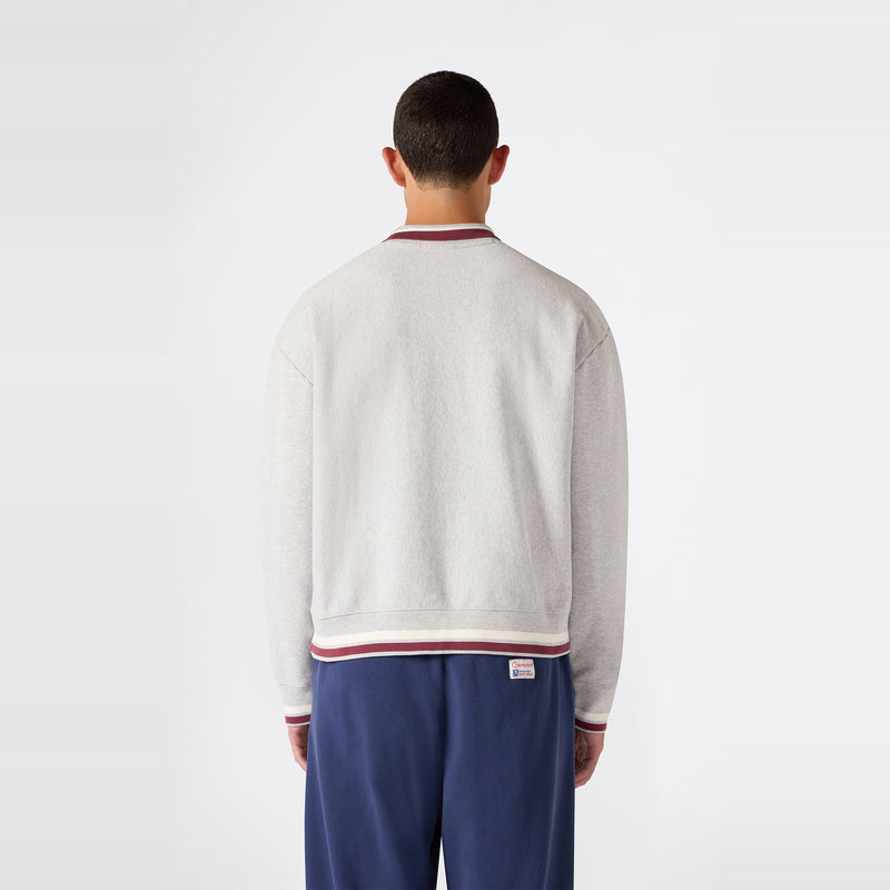 A person with short hair is standing with their back to the camera, wearing a light gray Champion Men’s Knitted Polo Neck Sweatshirt with maroon and white trim and blue pants. The background is plain white.