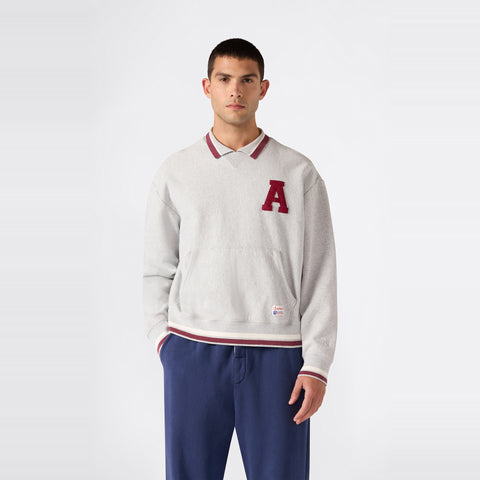 A man stands against a plain background, wearing a light gray Champion Men’s Knitted Polo Neck Sweatshirt featuring a red A on the left chest, layered over a collared shirt, and paired with blue pants.