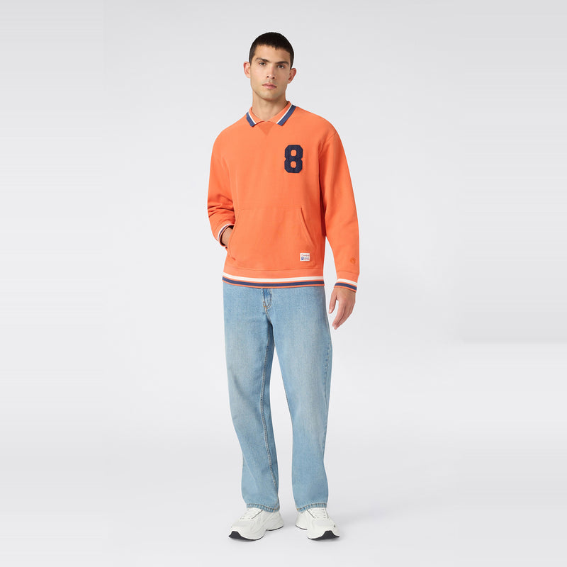 A man stands against a plain light gray background wearing the Champion Men’s Knitted Polo Neck Sweatshirt in vibrant orange, which features a navy 8 on the front, paired with light blue jeans and white sneakers. This men’s sportswear piece is crafted from soft Reverse Weave fleece for classic comfort and style.