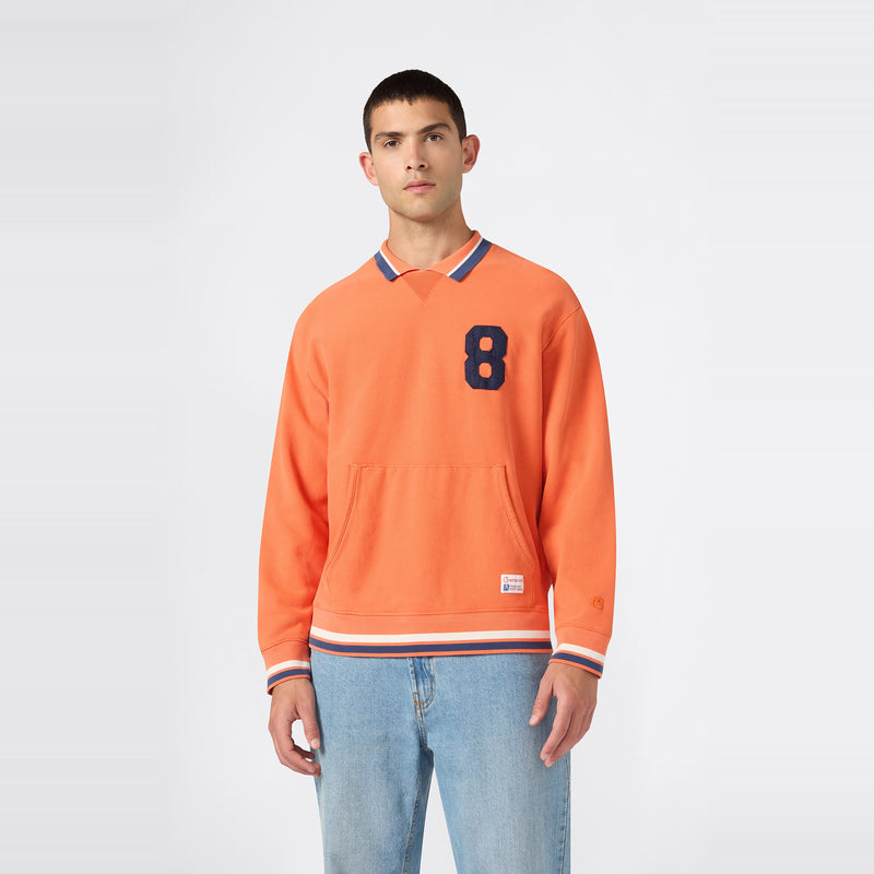 A man is wearing a Champion Men’s Knitted Polo Neck Sweatshirt in orange, featuring a blue number 8 on the chest, paired with light blue jeans, standing against a plain white background.